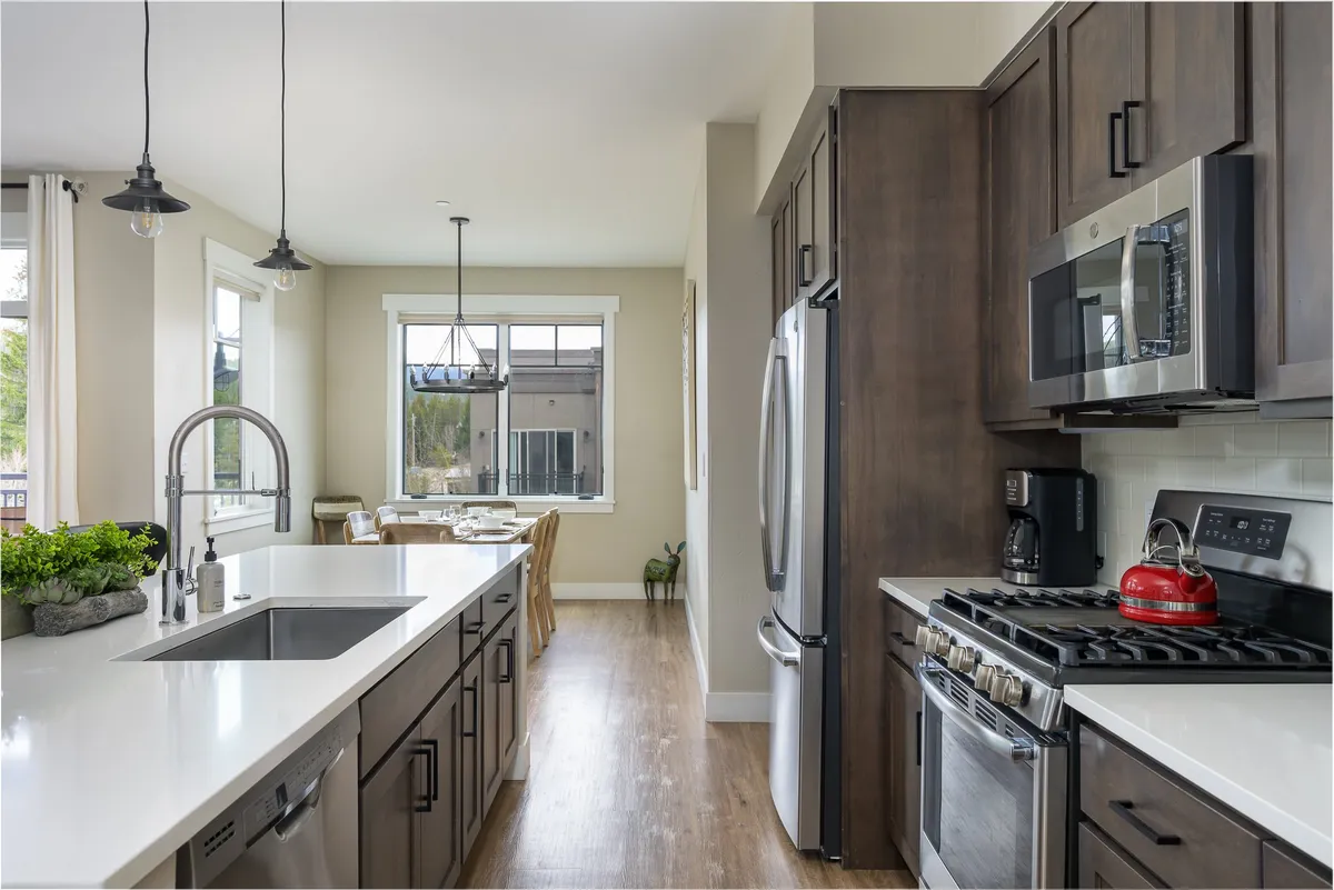 A spacious kitchen with a central island (including a sink), stainless steel appliances, and a stylish dining area visible through a large window. Decorative plants add a touch of warmth and homeliness, while the layout encourages group interaction and shared meals.