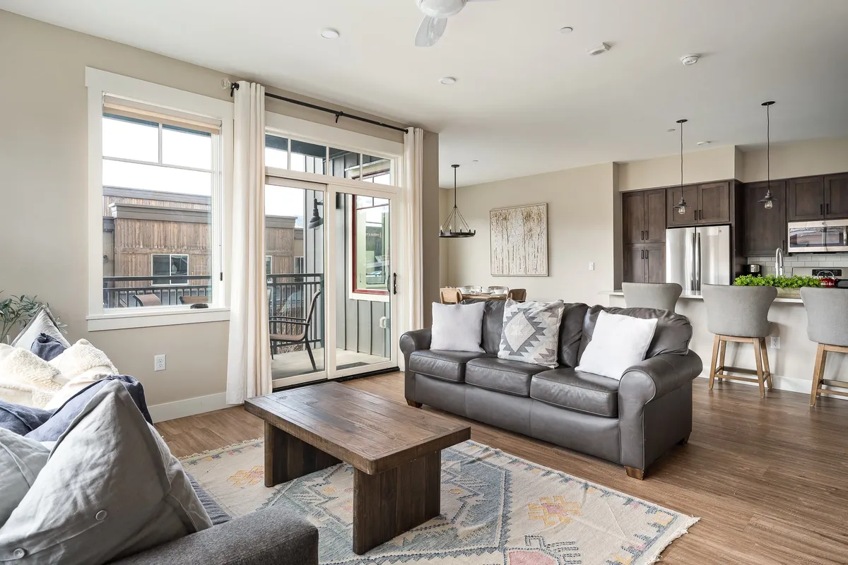 An open-concept interior with a comfortable sofa, coffee table, and bar stools at a breakfast bar. Balcony doors allow sunlight to pour in, hinting at an easy indoor-outdoor flow. The artwork, stylish furnishings, and visible balcony create a cohesive, welcoming social space.