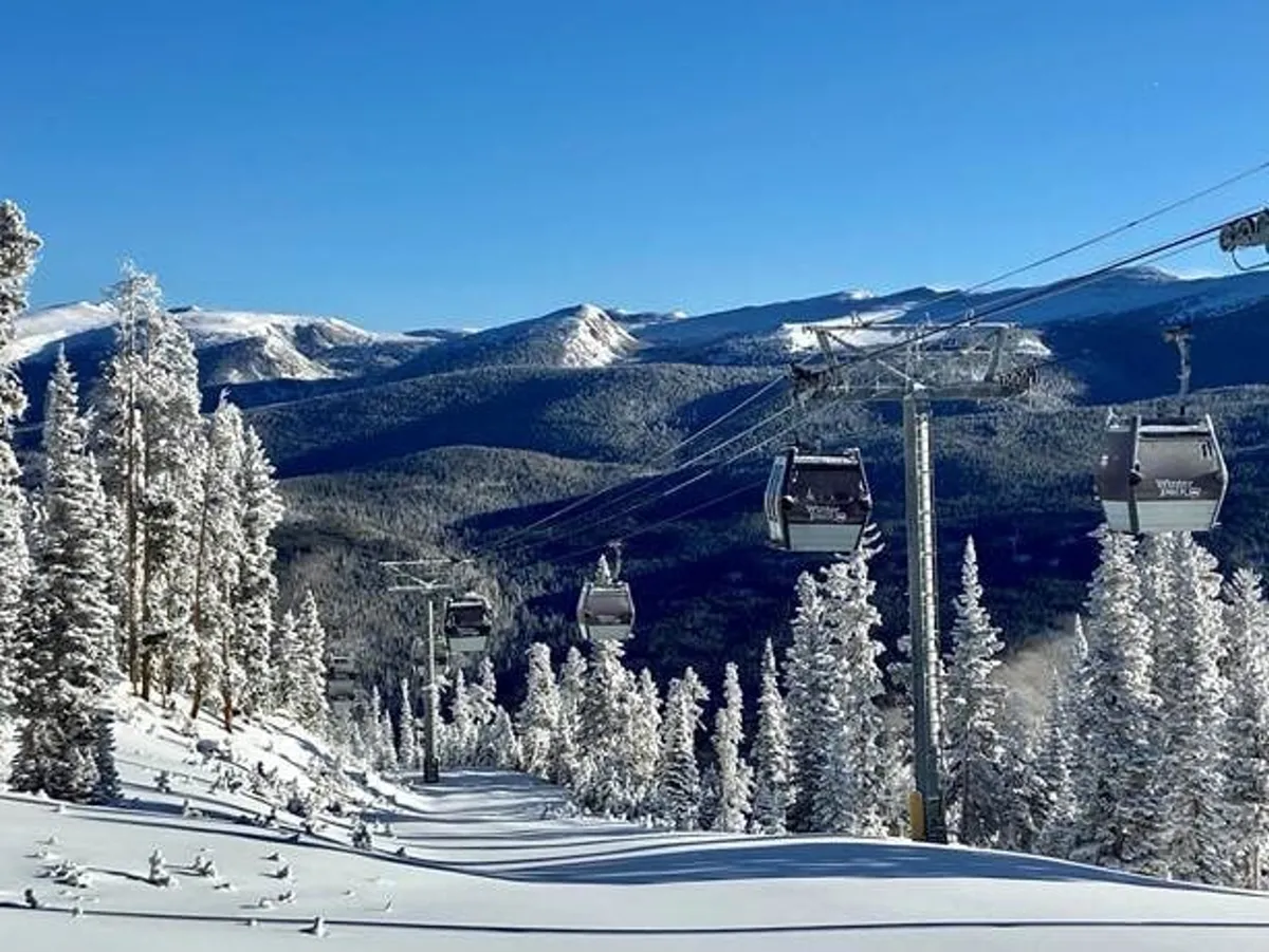 A snowy mountain landscape featuring ski lift gondolas ascending a snow-covered slope with distant mountains and evergreen trees. The scene captures the thrill and beauty of winter sports and mountain adventure in Winter Park.