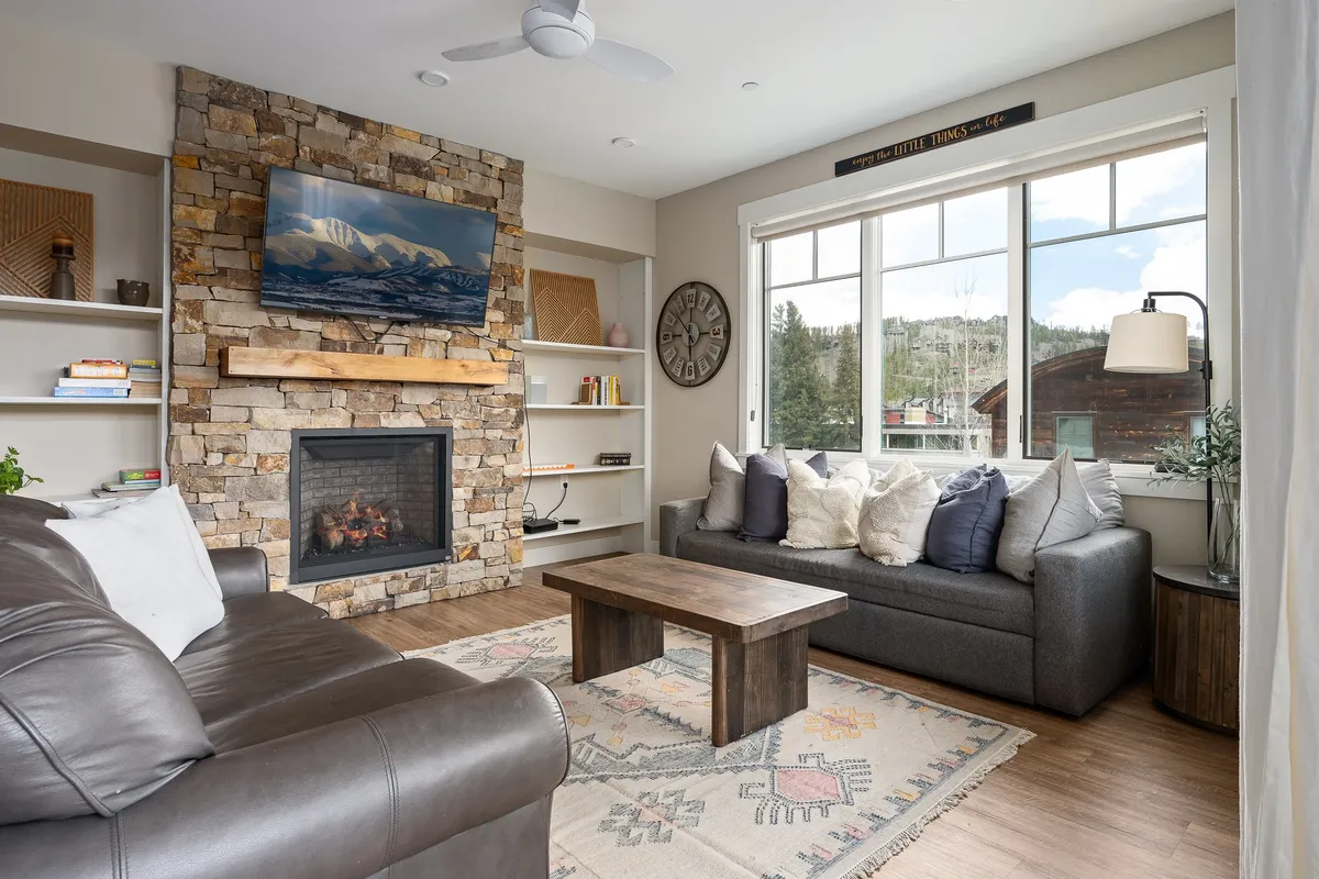 A warmly lit living room featuring a striking stone fireplace, large windows letting in natural light, comfortable sofas, and a central coffee table. The snow outside is visible through the expansive windows, reinforcing the mountain setting. The room exudes a sense of gathering and relaxation, ideal for cozy evenings or social moments.