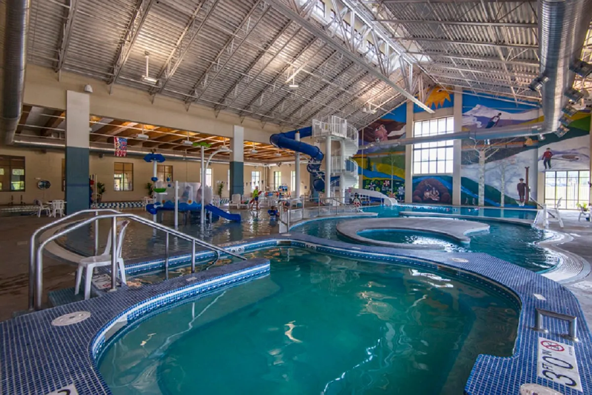 This energetic scene features a bright indoor swimming pool with a colorful water slide, playful murals, and water features. Families and children are seen enjoying the pool, capturing the fun, active recreation available nearby.