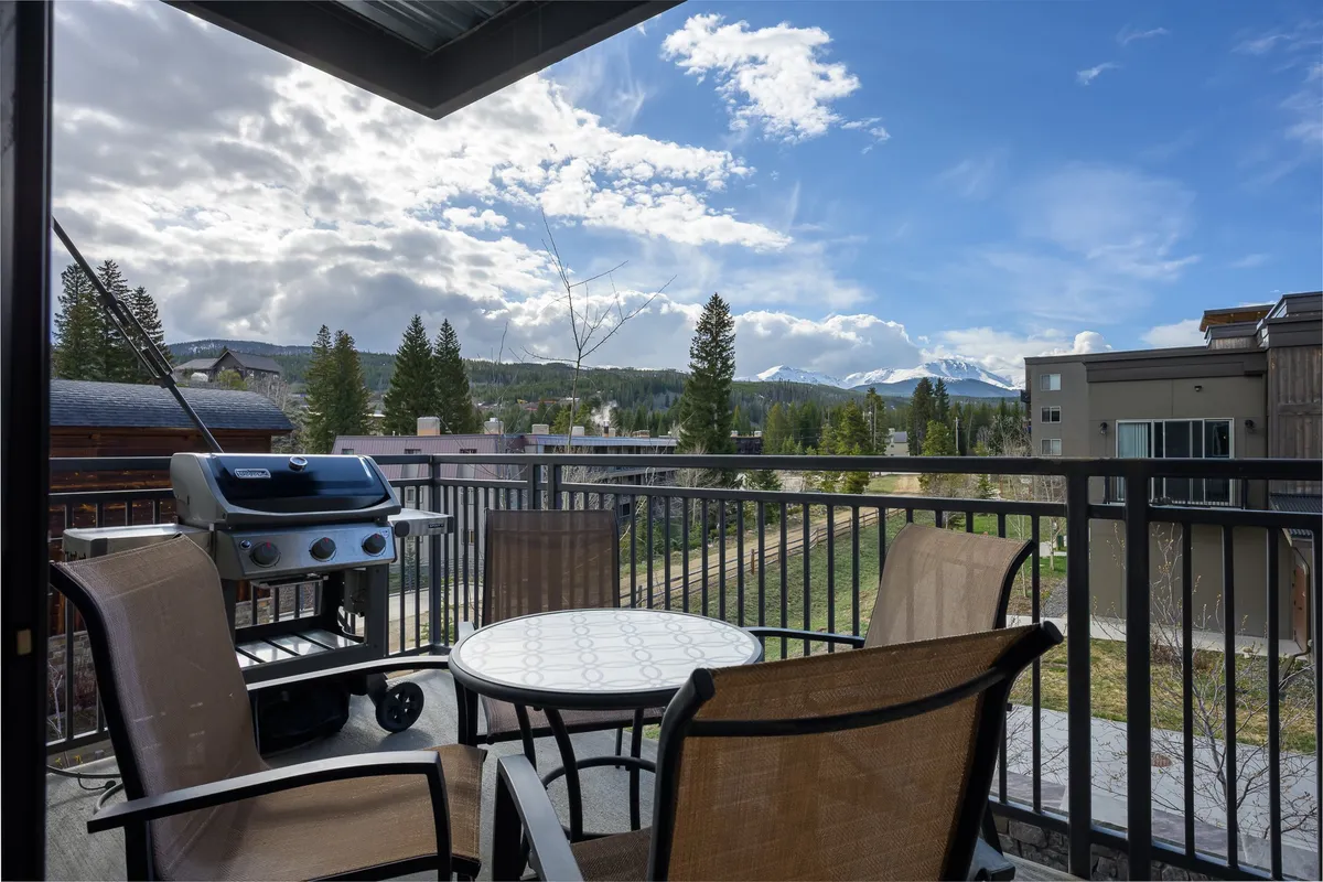 A private balcony setup featuring a grill, table, and chairs, all oriented to capture expansive mountain views. The scene suggests both relaxation and celebration, with the grill pointing to après-ski gatherings and the table set for outdoor dining against a stunning natural backdrop.