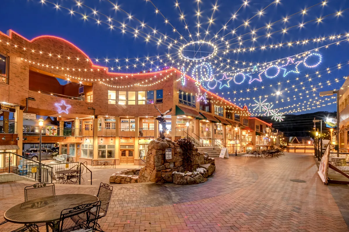 This image captures an outdoor scene with festive string lights, decorative stars, a stone fountain, and inviting outdoor seating areas. People can be seen gathering and enjoying the lively community atmosphere, suggesting a family-friendly environment and social opportunities.