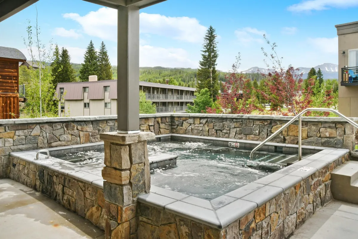 A tranquil outdoor jacuzzi set against a picturesque mountain backdrop, with steam rising from the hot water and lounge areas nearby. The surrounding landscape is visible, emphasizing year-round relaxation and the appeal of the community hot tub.