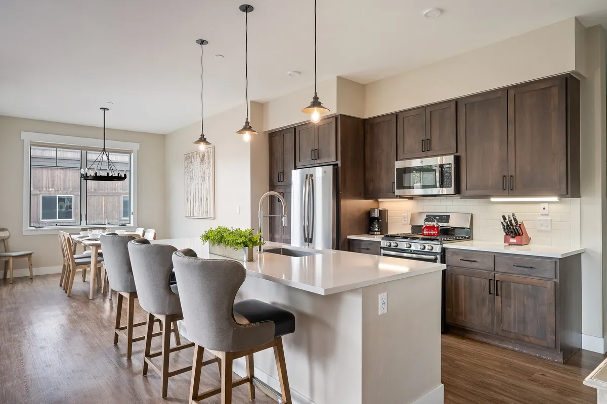 A bright and inviting kitchen scene featuring a kitchen island with bar stools, stainless steel appliances, and a dining area visible in the background. The space is styled for both functionality and social gatherings, with pendant lighting and modern décor.