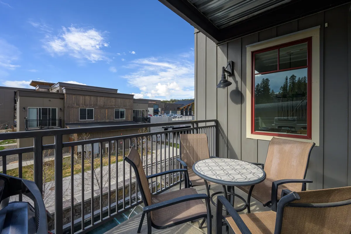 A peaceful balcony setup featuring a table with a patterned surface and two chairs, overlooking neighboring buildings and the surrounding landscape. The scene is set for relaxing with a morning coffee or sharing a quiet moment with friends while enjoying the fresh mountain air and scenic views.