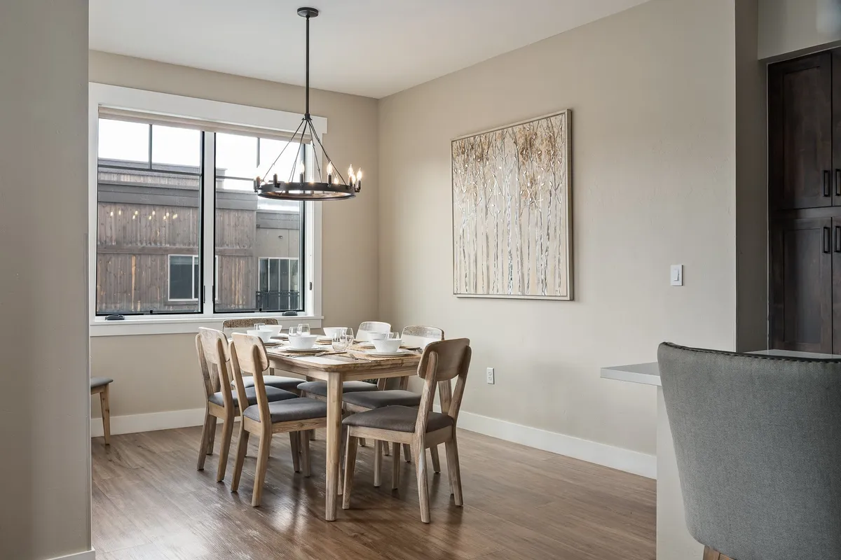 A stylish dining area featuring a table set beneath a chandelier, with artwork on the wall and a large window providing ample daylight. The space balances warmth, functionality, and a scenic vista ideal for casual work sessions or coffee breaks.