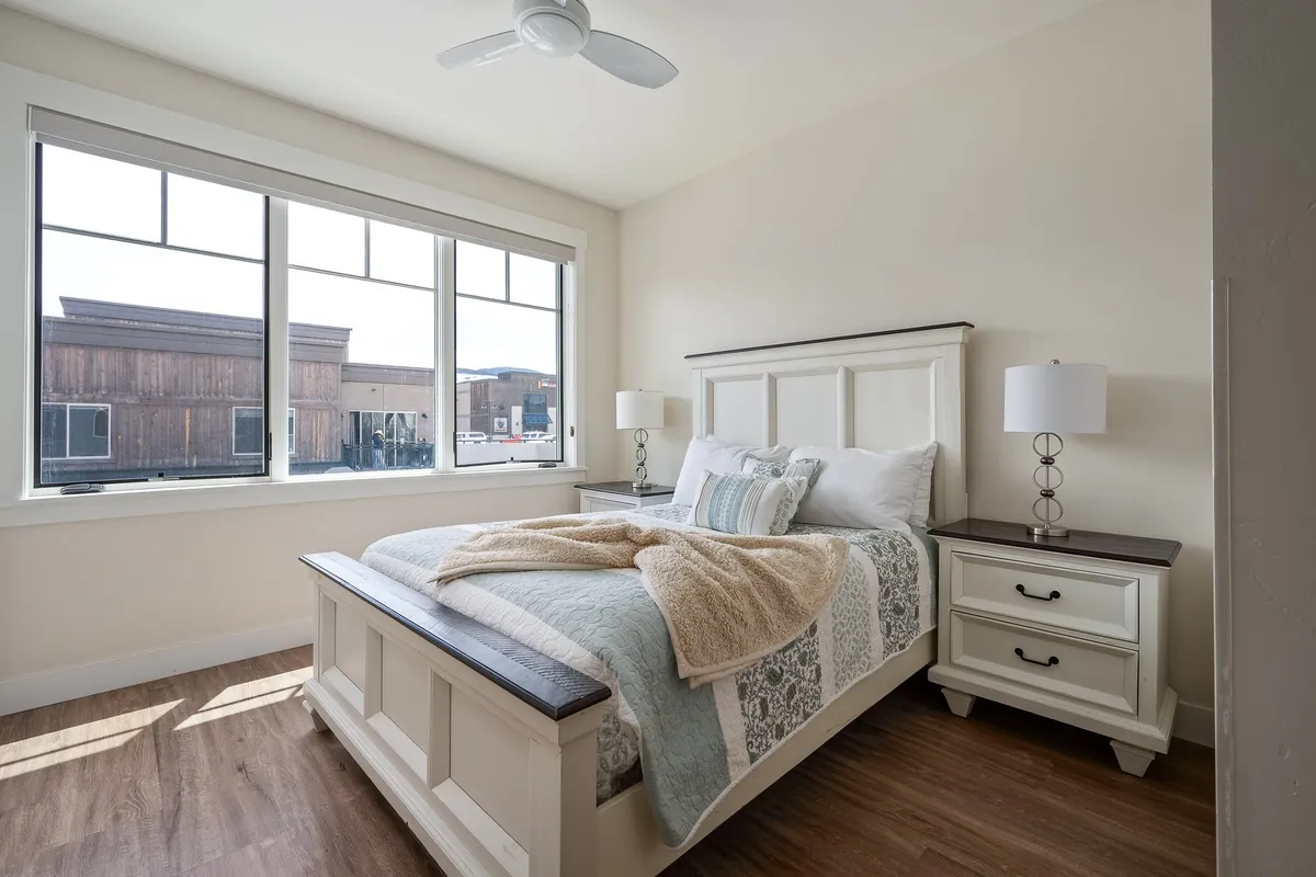 A serene second bedroom with a queen bed, decorative throw, large windows offering natural light and possible mountain views, and a nightstand with a lamp. The atmosphere is tranquil and inviting, ideal for guests seeking relaxation.