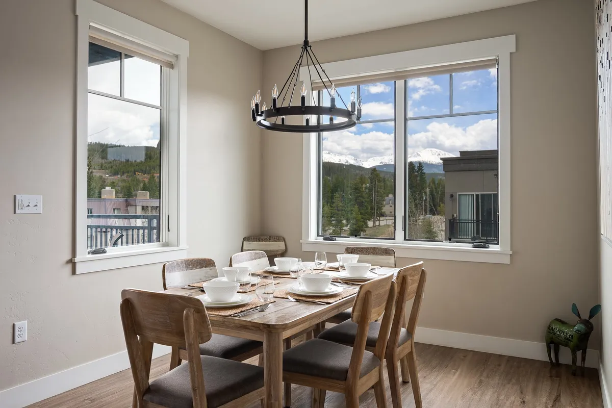 A cozy dining nook featuring a dining table set for six, a window view that fills the space with natural mountain light, and a modern chandelier overhead. The setting invites gatherings, family dinners, or board game nights.